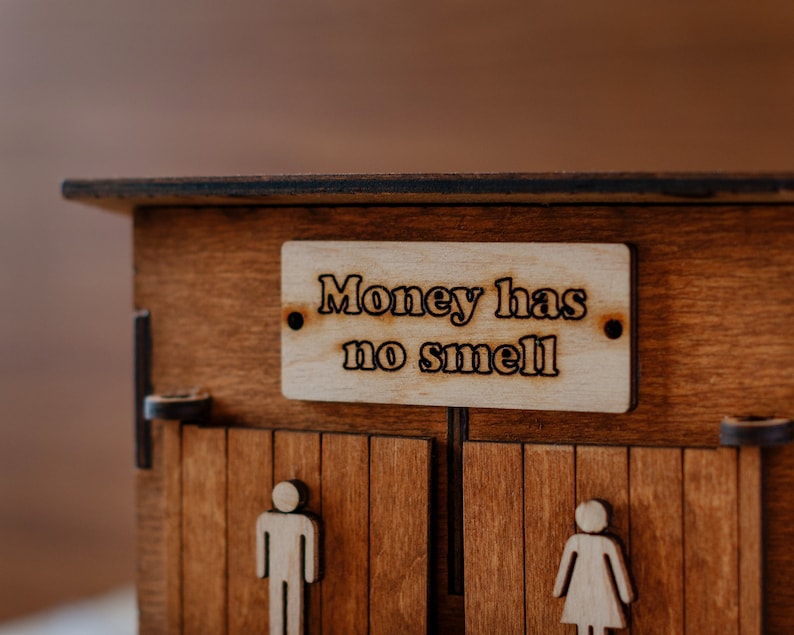 Puede incluir: Una caja de madera con dos puertas, cada una con la silueta de una persona. La caja tiene un letrero que dice "El dinero no huele."