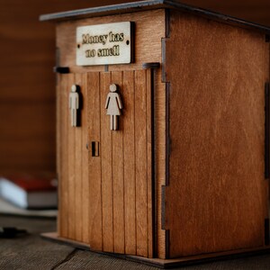 Puede incluir: Una caja de madera para guardar dinero con forma de letrina, con un letrero que dice "El dinero no huele".