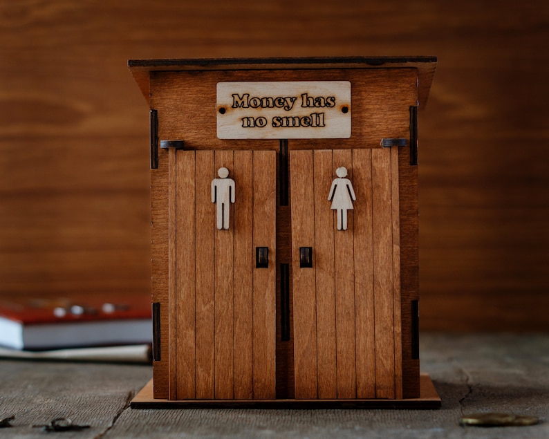 May include: Wooden outhouse-style money box with a sign that reads "Money has no smell". The box has two doors with male and female symbols.
