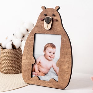 May include: Wooden bear-shaped picture frame with a photo of a baby. The frame is made of light brown wood and has a bear face design at the top. The baby is sitting on a gray blanket. A decorative basket with cotton balls is in the background.