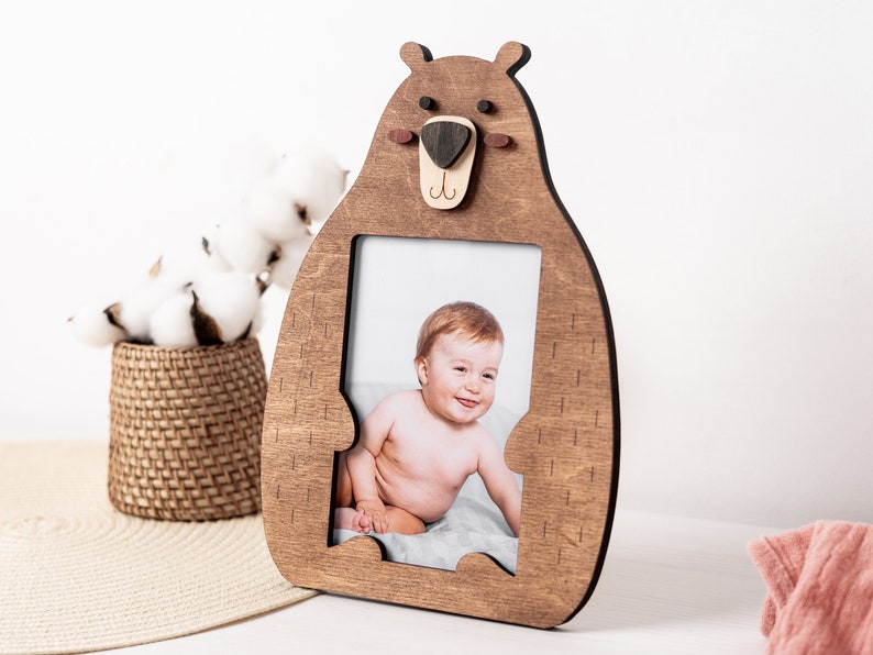 May include: Wooden bear-shaped picture frame with a photo of a baby. The frame is made of light brown wood and has a bear face design at the top. The baby is sitting on a gray blanket. A decorative basket with cotton balls is in the background.