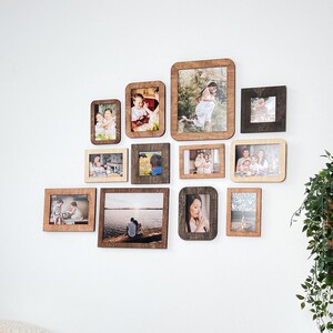 K&ouml;nnte beinhalten: Eine Ansammlung von Holzbilderrahmen in verschiedenen Gr&ouml;&szlig;en und Formen, die an einer wei&szlig;en Wand angebracht sind. Die Rahmen enthalten Fotos von Menschen und Landschaften. Die Rahmen sind in Braun- und Beiget&ouml;nen gehalten.