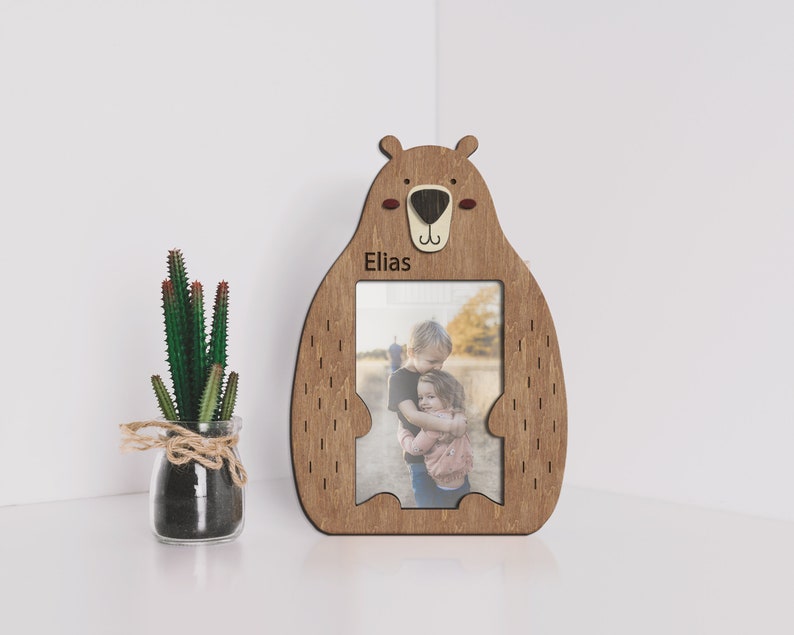 Wooden 4 by 6 inches photo frame in the shape of a cute sitting bear holding a photo. The name is engraved above the photo. This frame is 10 by 7 inches and 8 millimeters thick. The bear can be painted in different colors. The name can be changed.