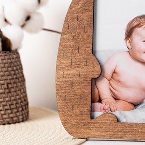 May include: Wooden photo frame with a unique, curved design. The frame is a warm brown color and holds a photograph of a smiling baby. The background includes a woven basket and cotton stems.