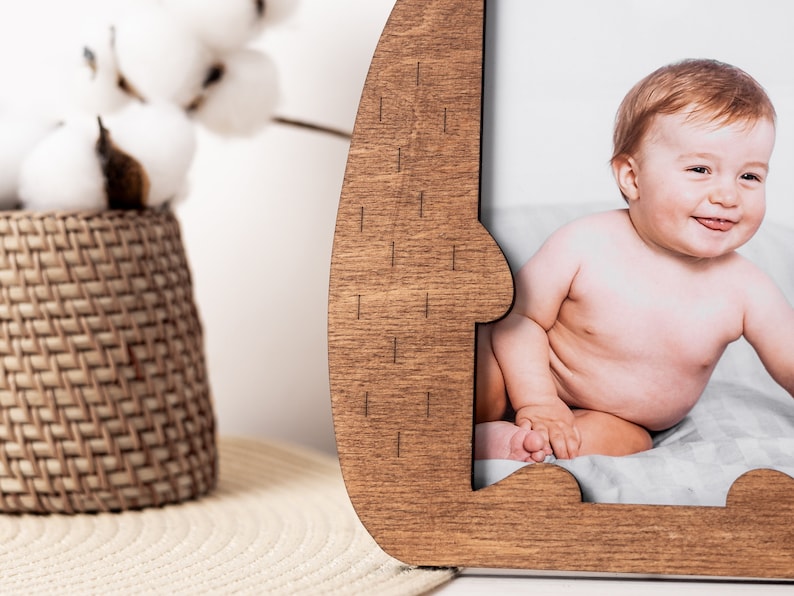 May include: Wooden photo frame with a unique, curved design. The frame is a warm brown color and holds a photograph of a smiling baby. The background includes a woven basket and cotton stems.