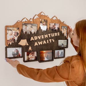 May include: A wooden photo frame with a mountain and forest design. The frame has multiple openings for photos and the text "Adventure Awaits" in the center. A person is hanging the frame on a white wall.