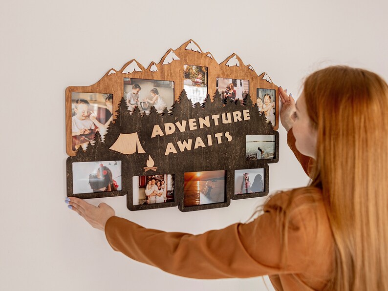 May include: A wooden photo frame with a mountain and forest design. The frame has multiple openings for photos and the text "Adventure Awaits" in the center. A person is hanging the frame on a white wall.