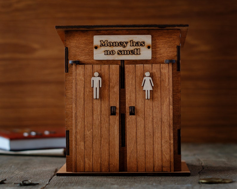 Puede incluir: Una caja de madera para guardar dinero con forma de letrina con dos puertas. El letrero sobre las puertas dice "El dinero no huele."