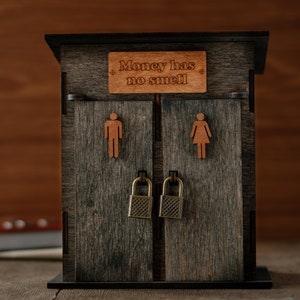 May include: A wooden outhouse-style box with a sign that reads "Money has no smell". The box has two doors with a lock on each door. The doors are closed and the box is sitting on a wooden surface.