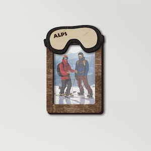 May include: A wooden photo frame with a ski goggle design. The frame has the word "ALPS" printed on it. The photo inside shows two people skiing in the snow.