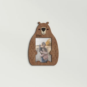 May include: Wooden bear-shaped picture frame with a photo of two children hugging. The frame is brown with a light brown nose and ears. The name "Elias" is printed on the frame. The frame is designed to hold a rectangular photo.