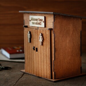 Puede incluir: Caja de madera para guardar dinero con forma de letrina con un letrero que dice "El dinero no huele".