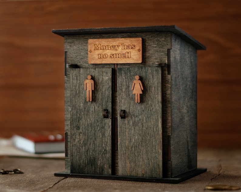 May include: A wooden outhouse-style money box with a sign that reads "Money has no smell". The box has two doors with a male and female symbol on each.