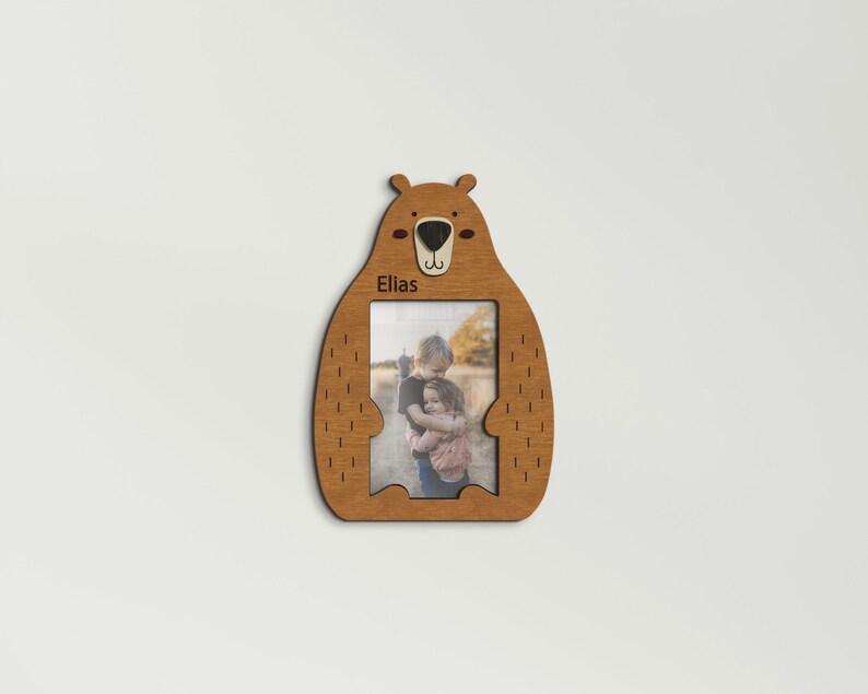 May include: Wooden bear-shaped picture frame with a photo of two children hugging. The frame is light brown with a bear face at the top and the name "Elias" printed on the frame. The frame is designed to hold a 4x6 inch photo.