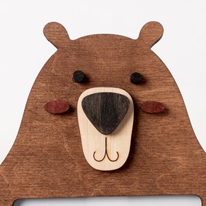 May include: Wooden bear-shaped wall decor with a dark brown finish. The bear's face features a light brown nose, black eyes, and red-brown cheeks. The design includes a cutout for a picture or other item.