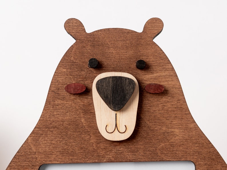 May include: Wooden bear-shaped wall decor with a dark brown finish. The bear's face features a light brown nose, black eyes, and red-brown cheeks. The design includes a cutout for a picture or other item.