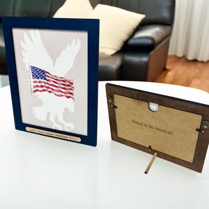 Puede incluir: Un marco azul marino con una impresión de un águila blanca sosteniendo una bandera estadounidense. La parte posterior del marco dice "Proud to be American".