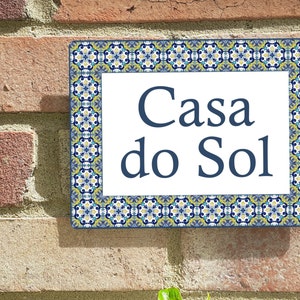 Pode incluir: Um sinal branco com texto azul que lê "Casa do Sol" rodeado por uma borda com um padrão azul, amarelo e branco.