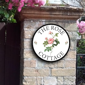Pode incluir: Um sinal redondo branco com letras pretas e uma borda preta. O sinal diz "The Rose Cottage" com uma rosa rosa e folhas verdes no centro.