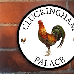 Op de afbeelding: Een wit rond bord met een zwarte rand en de tekst "Cluckingham Palace" in zwarte letters. Een kleurrijke haan staat in het midden van het bord met twee kleinere hanen aan weerszijden.