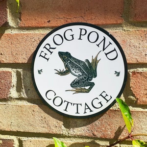 Può includere: Un cartello rotondo bianco con lettere nere che recita "Frog Pond Cottage". Una rana verde con macchie nere è al centro del cartello. Il cartello è montato su un muro di mattoni.