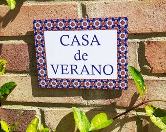 May include: A white sign with blue lettering that reads "CASA de VERANO" against a brick wall. The sign has a blue and red patterned border.
