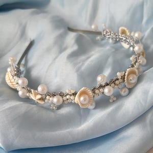 Può includere: Una fascia per capelli argentata adornata con perle finte e dettagli floreali color crema. Il design delicato presenta perle di varie dimensioni e piccoli abbellimenti a forma di fiore, creando un accessorio elegante. La fascia poggia su un tessuto azzurro.