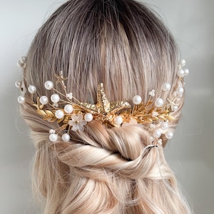 Könnte beinhalten: Ein goldfarbenes Haaraccessoire mit Seestern, Perlen und floralen Details. Das Haarteil ist vor blondem Haar platziert, das in lockeren Wellen und einem gedrehten Half-Updo gestylt ist. Dieses elegante Haaraccessoire ist für Hochzeiten geeignet.