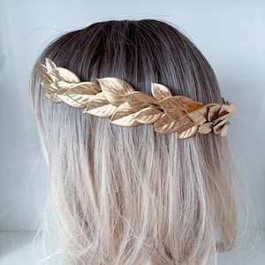 May include: A gold leaf crown headband with a simple design. The headband is made of metal and has a series of leaves that are connected together.