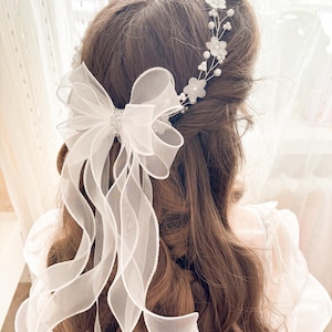 May include: A white bridal hair accessory featuring a large bow with long, flowing ribbons and a sparkling rhinestone center. Delicate floral and pearl accents adorn the hairpiece, creating an elegant and romantic look for weddings or special occasions.