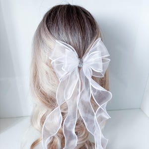 Könnte beinhalten: Eine weiße Organza-Haarschleife mit einem funkelnden Strass-Mittelteil. Die Schleife hat lange, fließende Bänder, die nach unten fallen. Das Haaraccessoire ist vor einem Hintergrund aus blondem und braunem Haar angebracht.