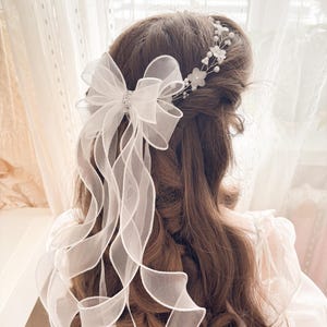 May include: A white organza bow with long, flowing ribbons and a sparkling center adorns the back of the head. A delicate floral and pearl hairpiece is also visible. The hair is long and brown, with soft curls.
