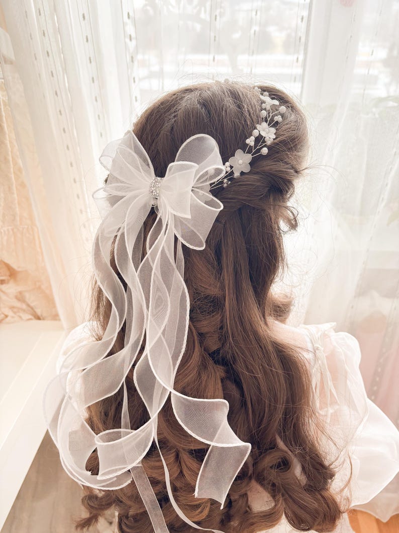 May include: A white organza bow with long, flowing ribbons and a sparkling center adorns the back of the head. A delicate floral and pearl hairpiece is also visible. The hair is long and brown, with soft curls.