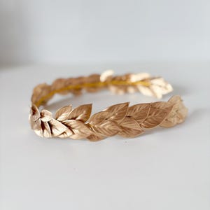 May include: A gold laurel wreath headband with a simple design. The headband is made of metal and has a series of leaves that are connected together.