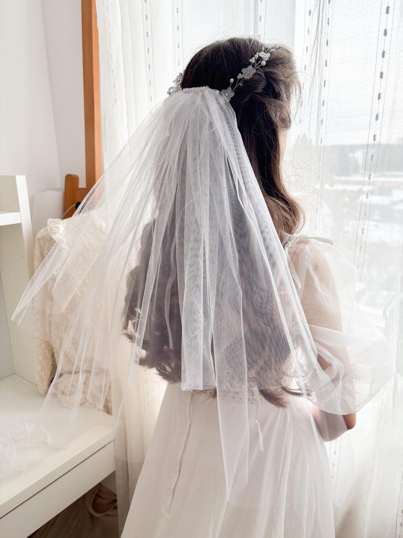 May include: A sheer white bridal veil with a delicate floral headpiece. The veil is layered and drapes over the back of a white dress. The image is taken indoors with natural light.