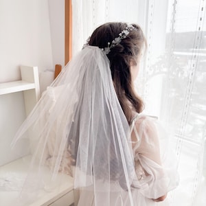 May include: A white tulle veil and floral headband. The veil is layered and drapes over the shoulders. The headband features small white flowers and pearls. The person is wearing a white dress with sheer sleeves, standing near a window with white curtains.
