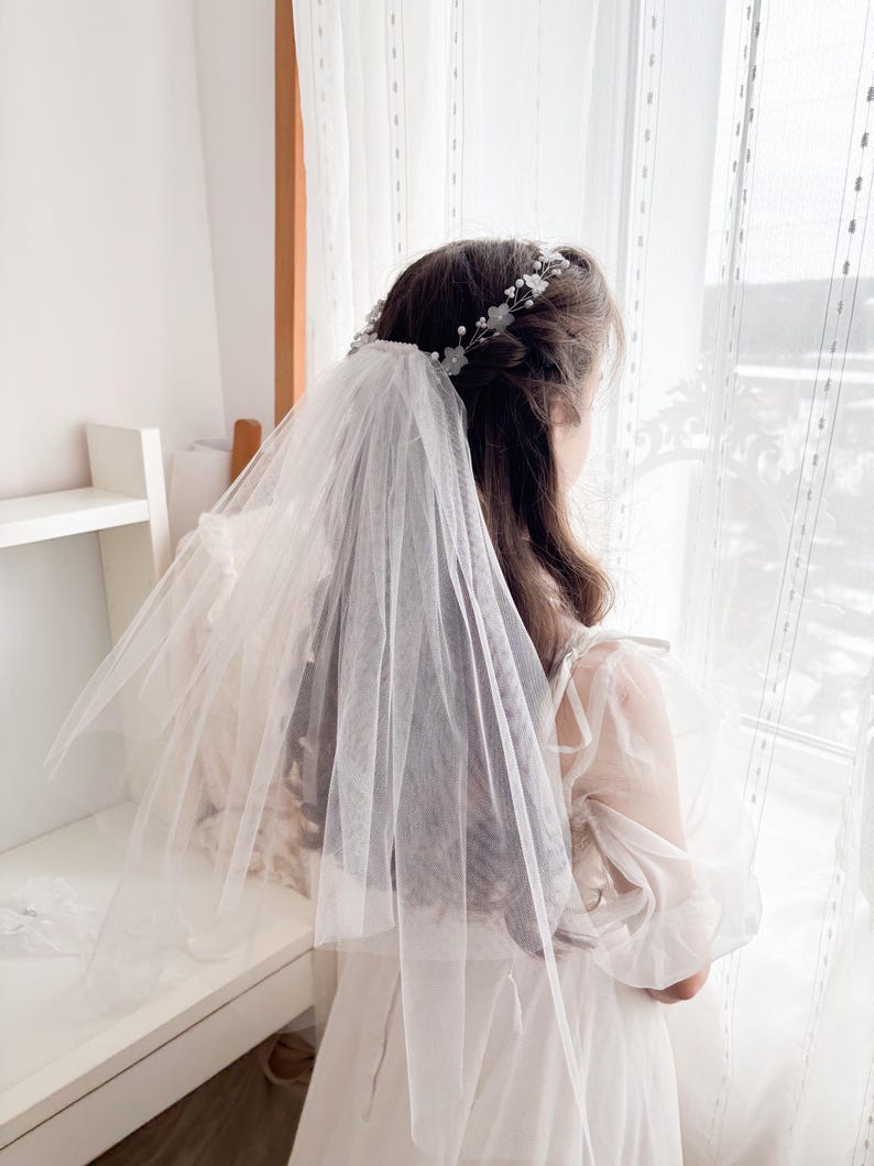 May include: A white tulle veil and floral headband. The veil is layered and drapes over the shoulders. The headband features small white flowers and pearls. The person is wearing a white dress with sheer sleeves, standing near a window with white curtains.