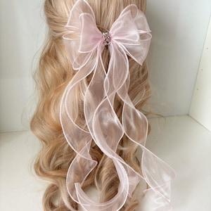 Puede incluir: Un lazo para el pelo de organza rosa claro con largas cintas onduladas. El lazo está asegurado con un pequeño adorno brillante. El accesorio para el cabello se coloca sobre un fondo de cabello rubio ondulado.