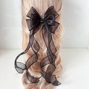 Puede incluir: Un lazo para el cabello de organza negra con un centro de satén y largas cintas fluidas. El lazo está sujeto a una pinza para el cabello y se muestra en el cabello de una persona con cabello rubio. Un accesorio para el cabello elegante.