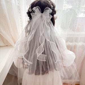 May include: A white bridal veil with a large bow and a sparkling embellishment. The veil has a layered design with delicate, wavy edges. A floral hairpiece is also visible, adding to the elegant aesthetic. The veil is designed for a wedding or special occasion.
