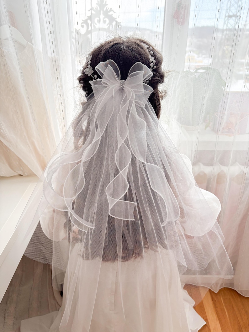May include: A white bridal veil with a large bow and a sparkling embellishment. The veil has a layered design with delicate, wavy edges. A floral hairpiece is also visible, adding to the elegant aesthetic. The veil is designed for a wedding or special occasion.