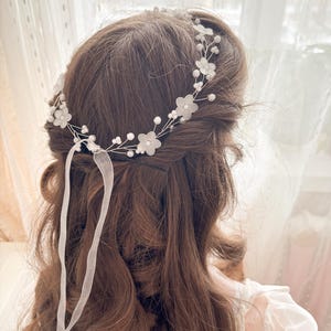 May include: A delicate bridal hairpiece featuring a floral design with small white flowers and faux pearls. The headpiece is made of silver wire and includes white ribbon streamers. The hair accessory is designed for a wedding or special occasion.