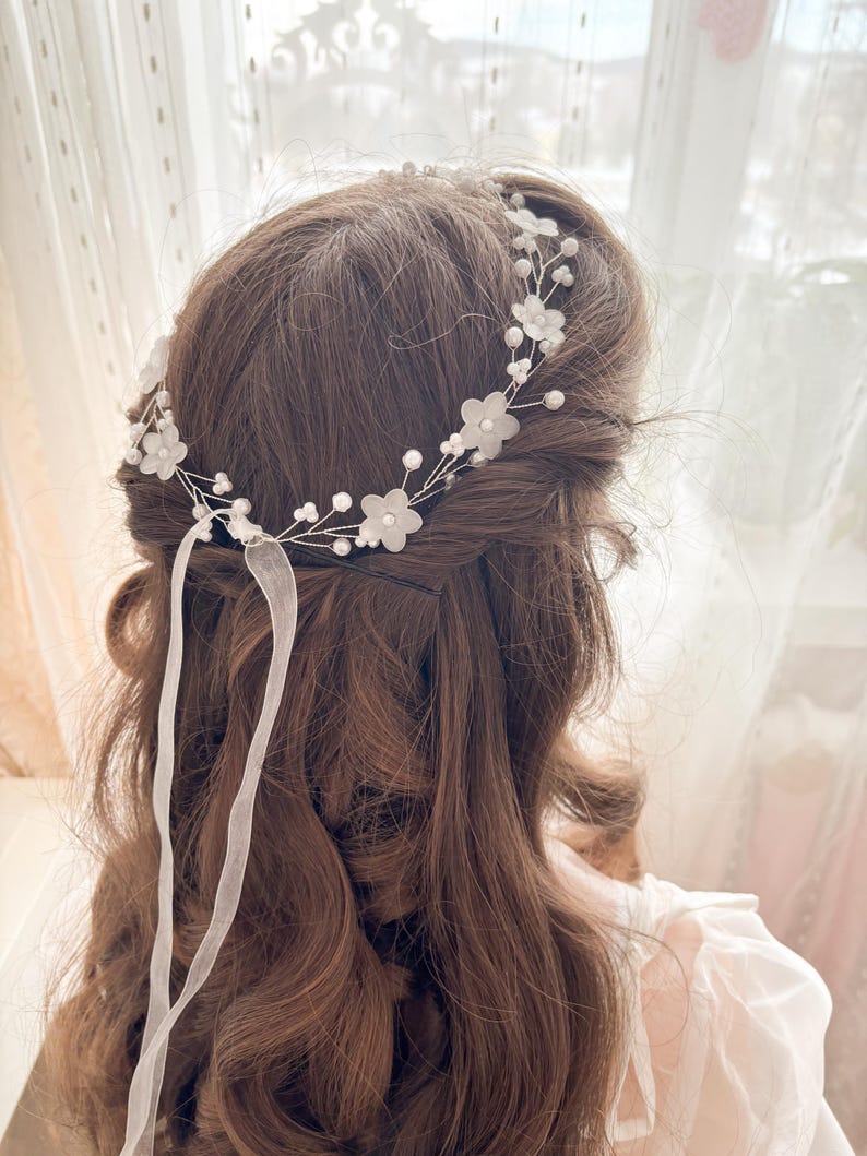 May include: A delicate bridal hairpiece featuring a floral design with small white flowers and faux pearls. The headpiece is made of silver wire and includes white ribbon streamers. The hair accessory is designed for a wedding or special occasion.
