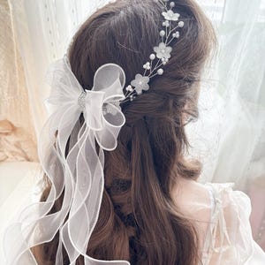 May include: A white bridal hair accessory featuring a delicate floral headband and a sheer bow with flowing ribbons. The headband has small white flowers and pearls. The bow has a sparkling rhinestone center.