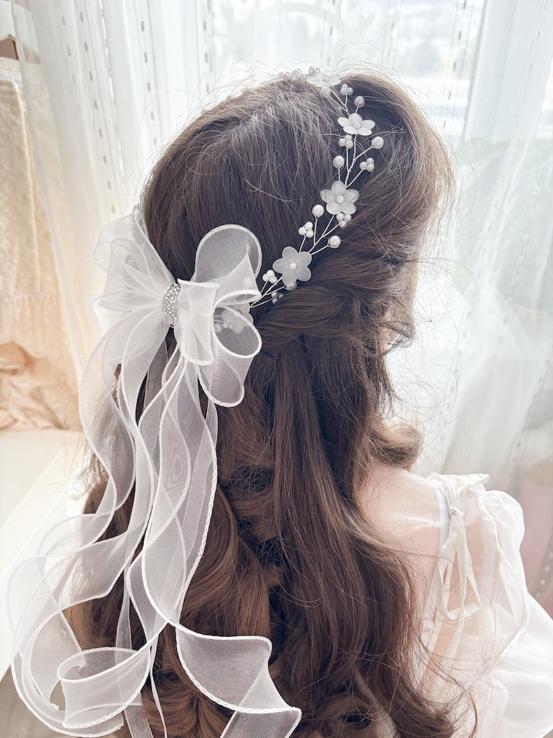 May include: A white bridal hair accessory featuring a delicate floral headband and a sheer bow with flowing ribbons. The headband has small white flowers and pearls. The bow has a sparkling rhinestone center.
