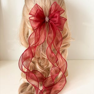 May include: A burgundy hair bow with a sparkling rhinestone centre. The bow is made of sheer, ruffled fabric and has long, flowing ribbons. The hair accessory is set against a backdrop of blonde hair.