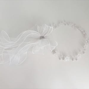 May include: A white bridal headpiece with a sheer veil and a floral crown. The veil features a large bow with a sparkling rhinestone accent. The floral crown is adorned with small white flowers and pearls, creating an elegant look.