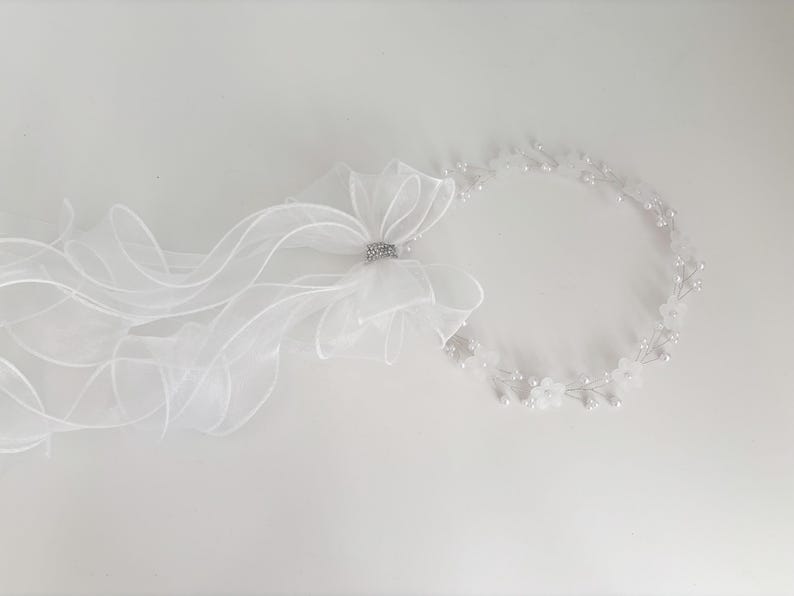 May include: A white bridal headpiece with a sheer veil and a floral crown. The veil features a large bow with a sparkling rhinestone accent. The floral crown is adorned with small white flowers and pearls, creating an elegant look.