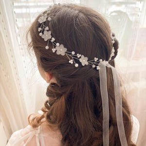 May include: A delicate bridal hairpiece featuring white fabric flowers and small pearl accents. The floral design is set on a silver wire frame, with sheer white ribbons cascading down the back. The hair accessory is designed for a special occasion.