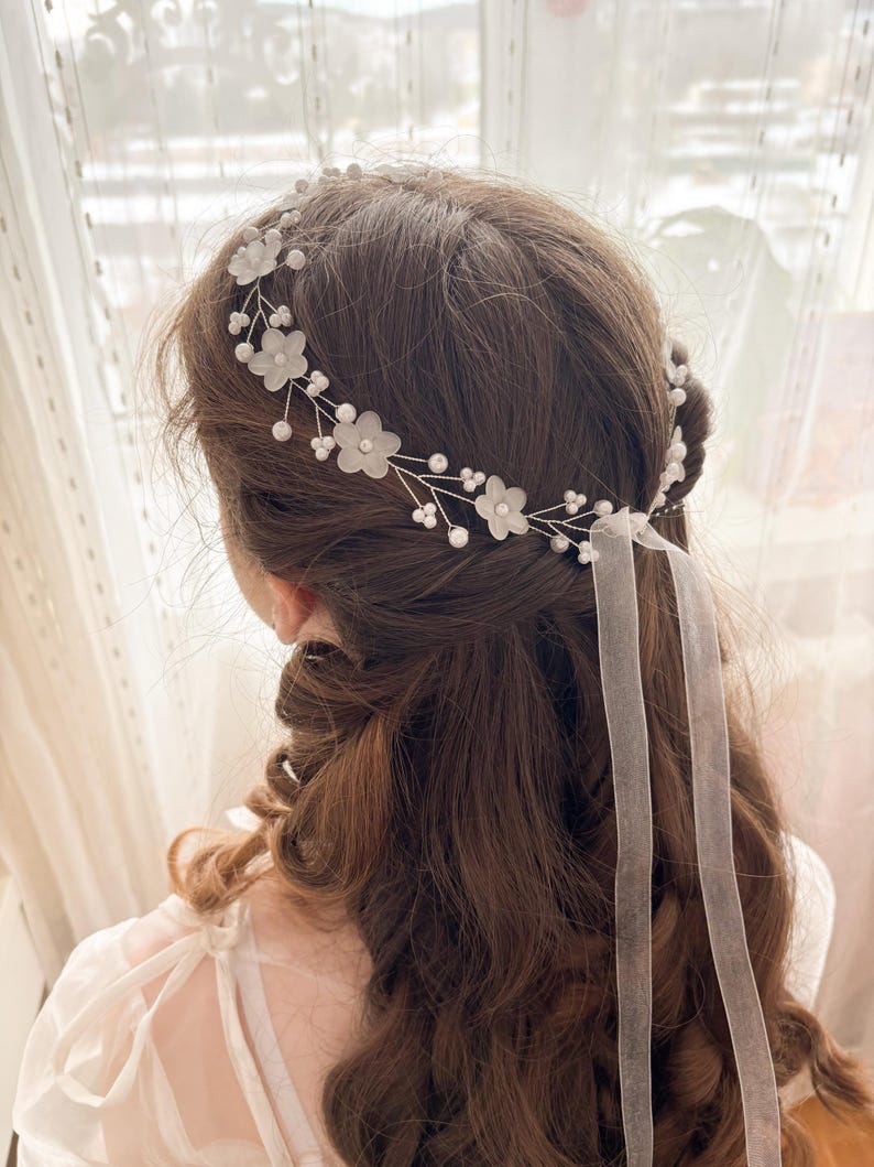 May include: A delicate bridal hairpiece featuring white fabric flowers and small pearl accents. The floral design is set on a silver wire frame, with sheer white ribbons cascading down the back. The hair accessory is designed for a special occasion.